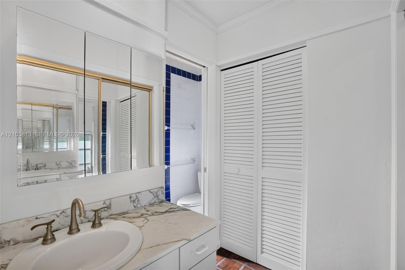 Vanity featuring a stone-patterned countertop, oval undermount sink, and multi-panel medicine cabinet with brass trim, complemented by white louvered closet doors