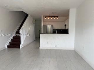 Open concept interior featuring wood-finish flooring, staircase with white railing, kitchen pass-through counter, stainless steel refrigerator, and industrial-style multi-light fixture