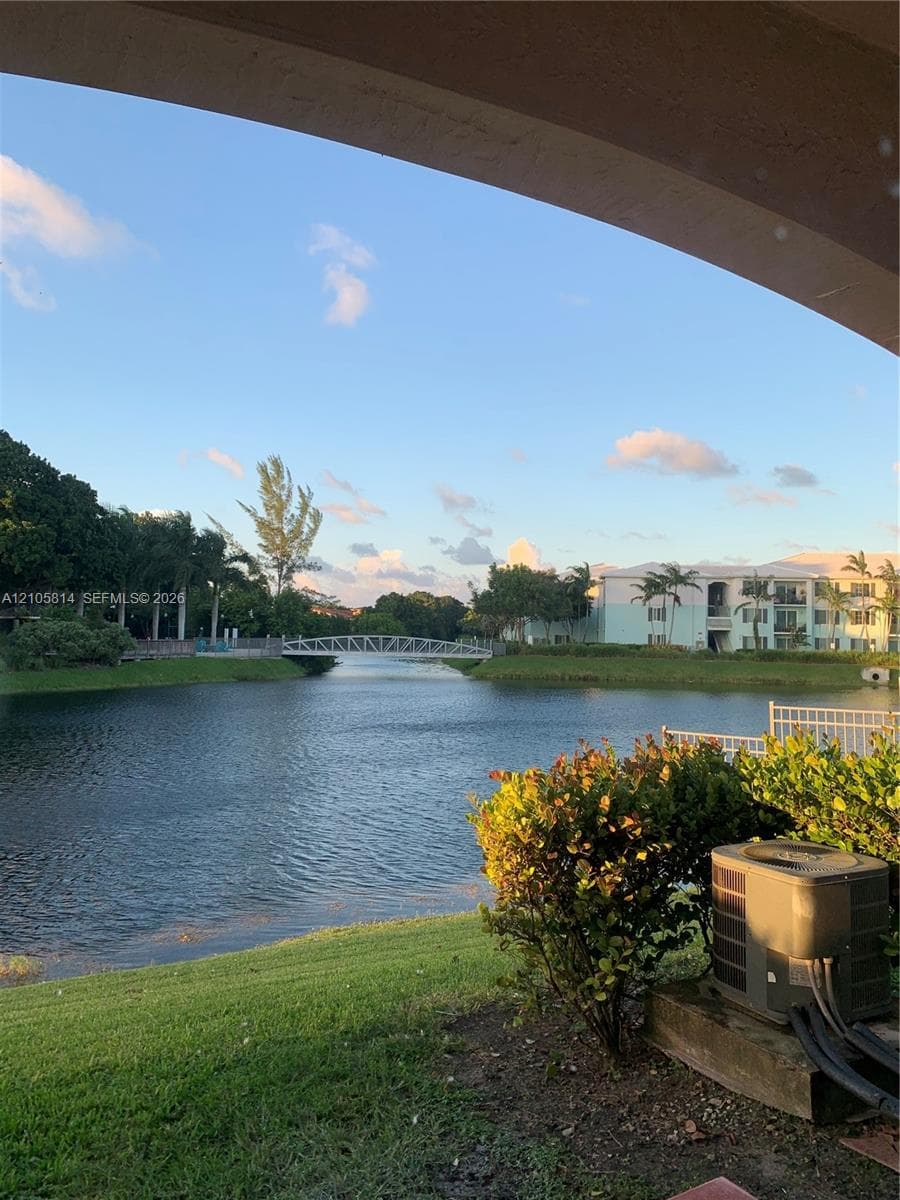 Waterfront property featuring a manicured lawn, lake, pedestrian bridge, and established landscaping