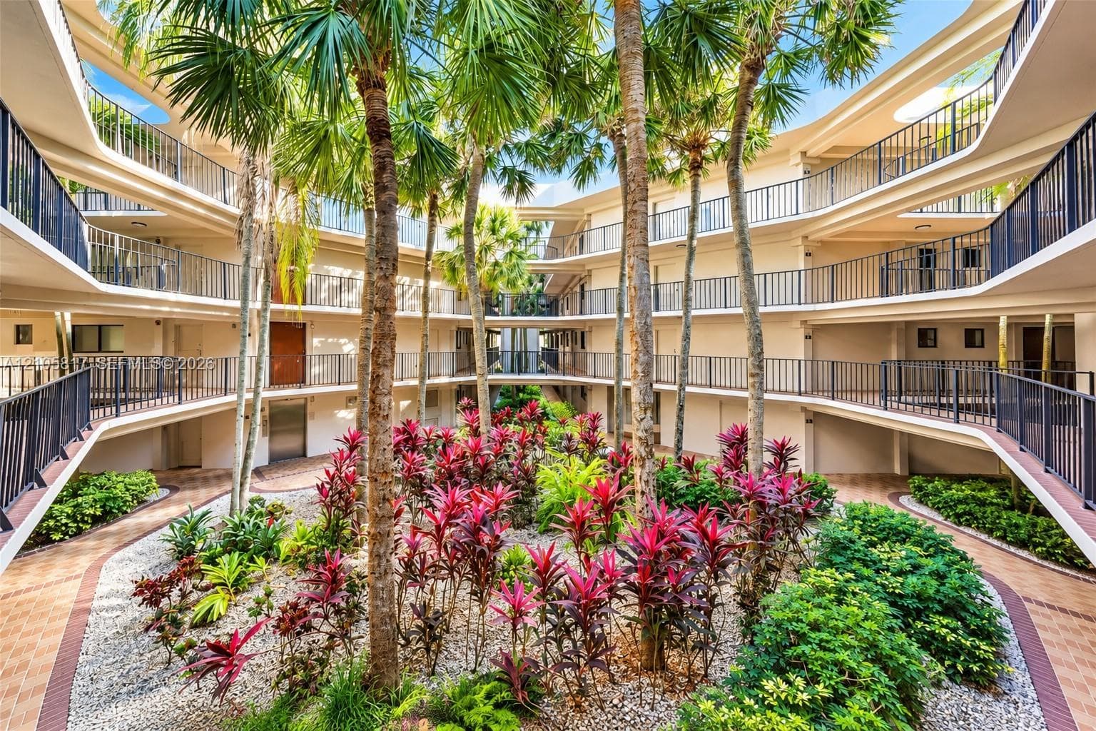 Central atrium courtyard featuring multi-level corridors with dark metal railings, mature palm trees, landscaped garden beds with tropical foliage, and paved walkways