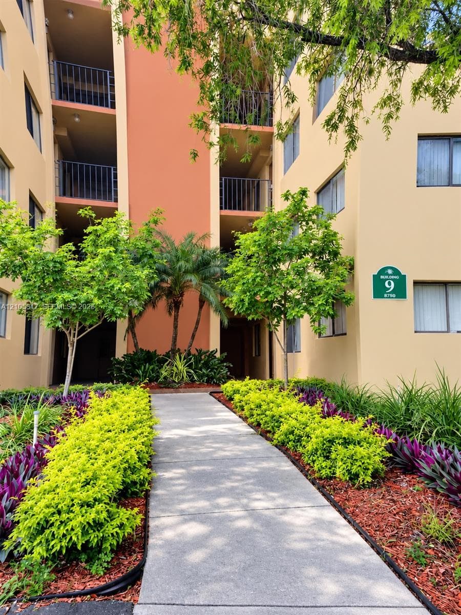 Multi-story exterior featuring stucco siding, integrated balconies with metal railings, a concrete walkway, and landscaped garden beds with palm trees