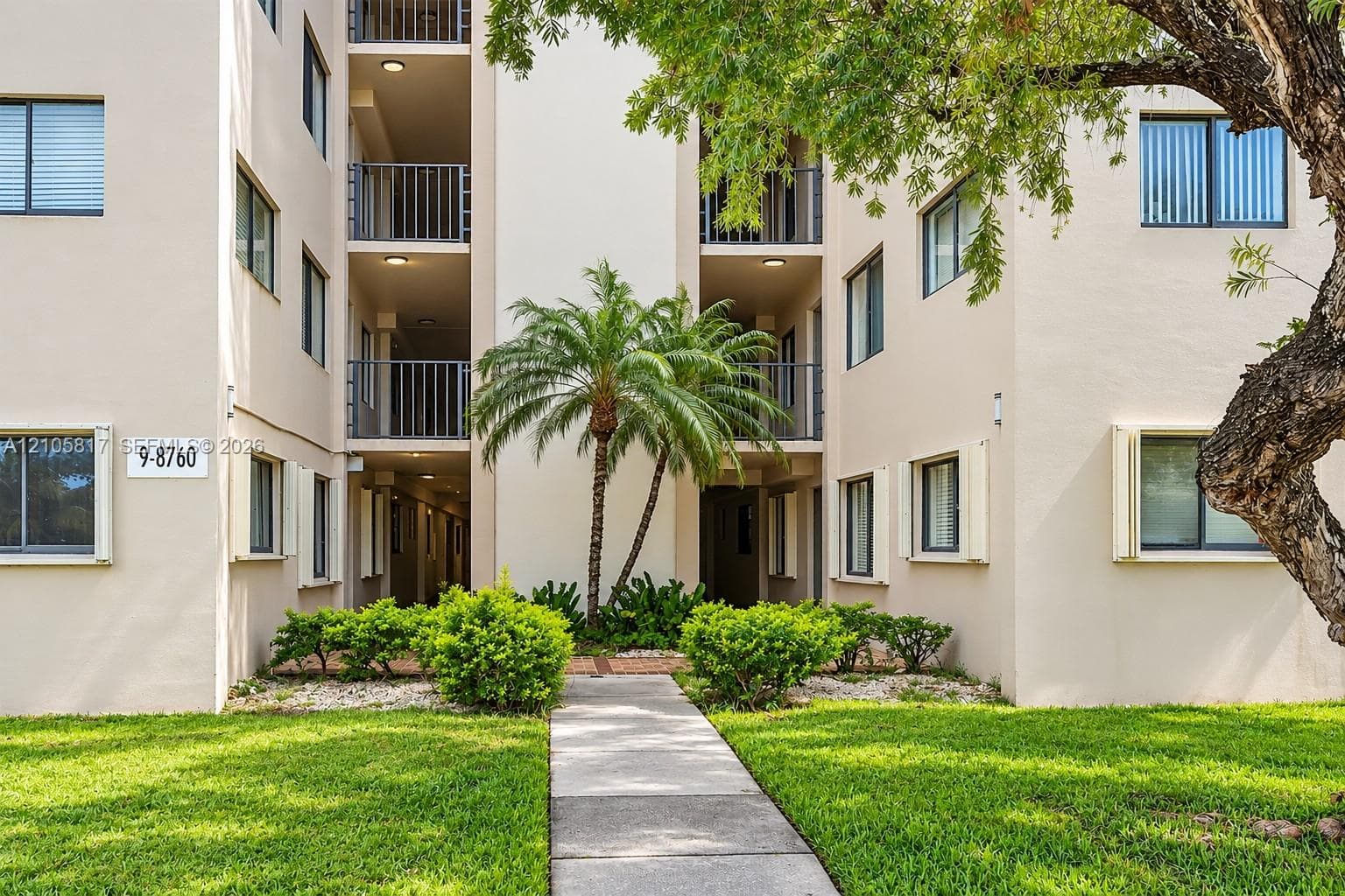 Multi-story stucco exterior featuring stacked balconies with metal railings, a central paved walkway, mature palm trees, and manicured landscaping