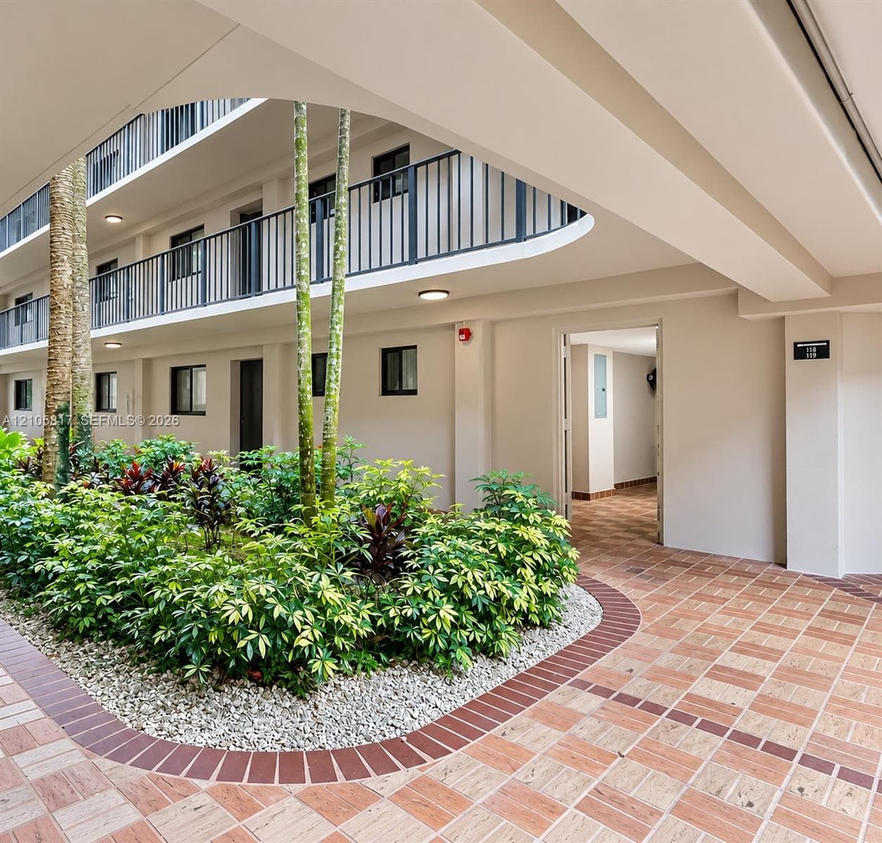 Multi-story architecture with arched structural detailing, stone-tiled pedestrian pathways, metal balcony railings, and manicured central landscaping with mature palms