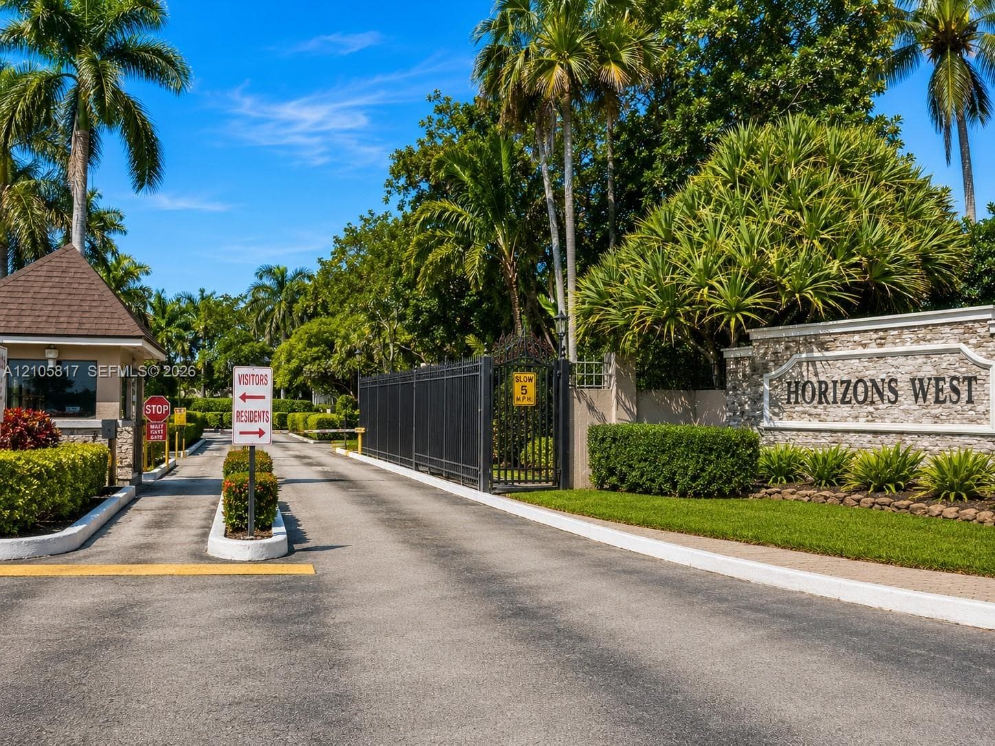Gated community entrance featuring a stone-clad monument sign, iron security gate, guardhouse, and paved access lanes