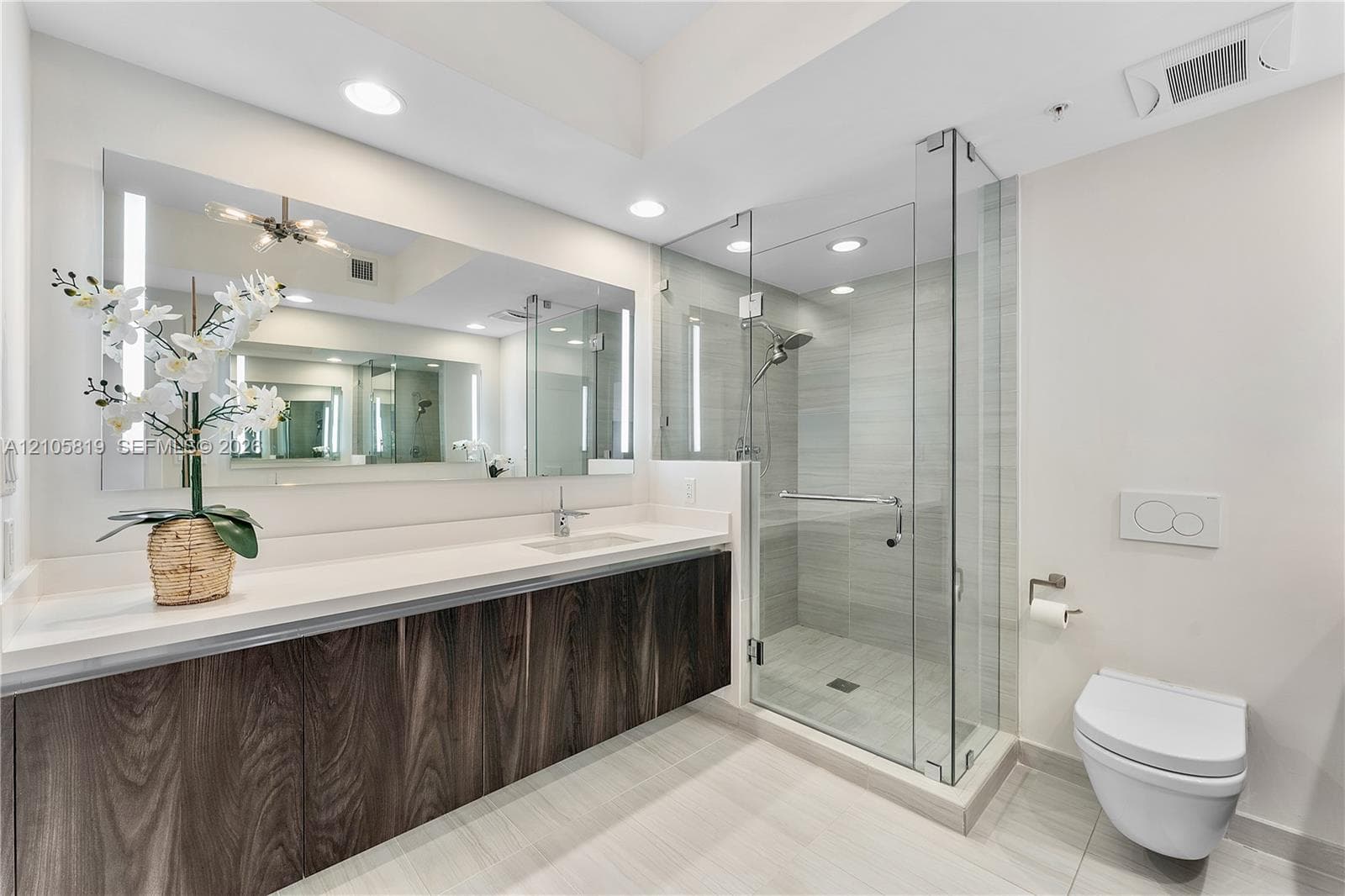 Floating vanity with wood-finish cabinetry, solid surface countertop, expansive wall-mounted mirror, glass-enclosed shower with tiled surround, and wall-mounted toilet