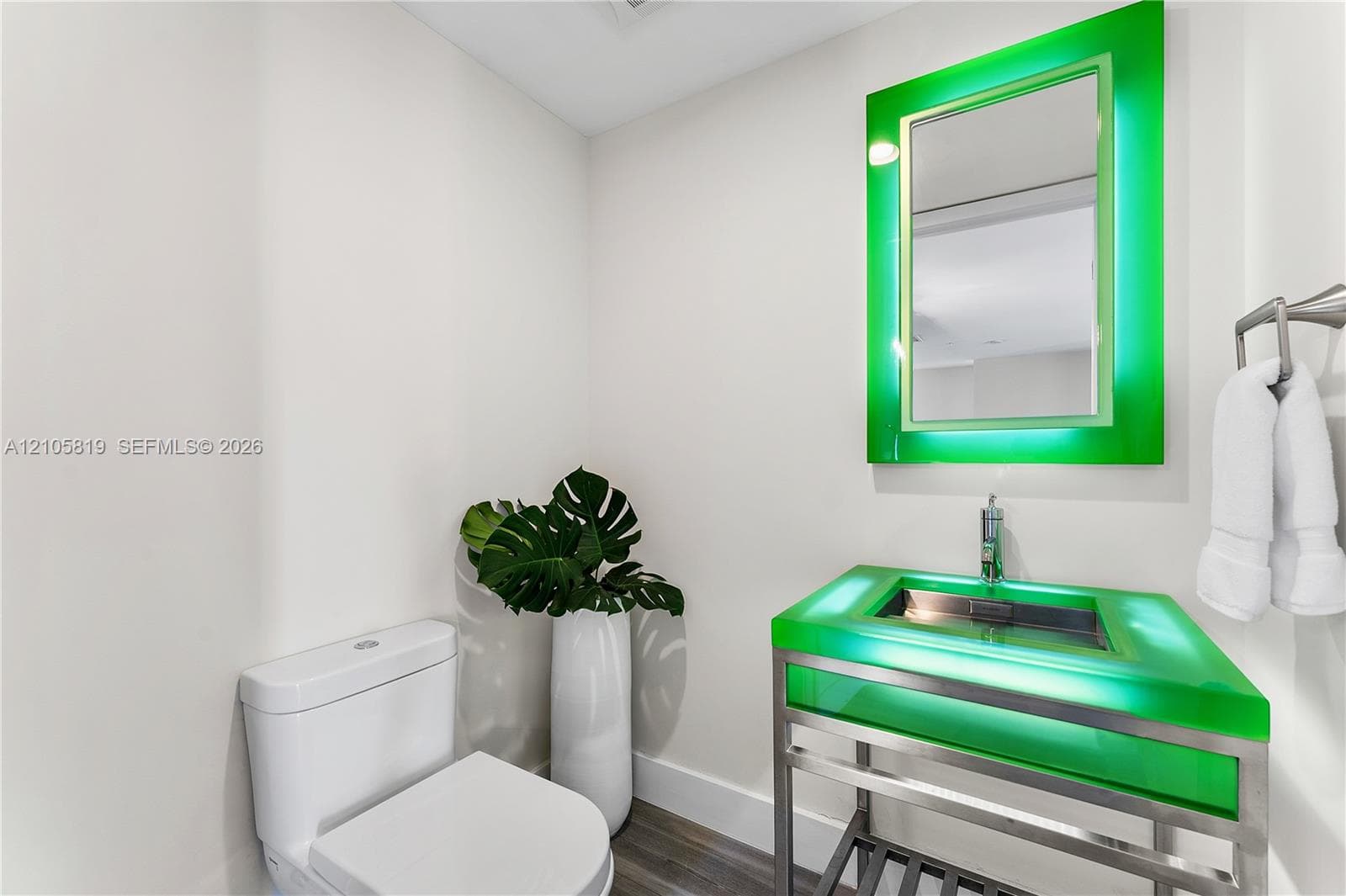 Floating vanity with an integrated green glass sink, polished metal frame, matching illuminated mirror, and wood-finish flooring