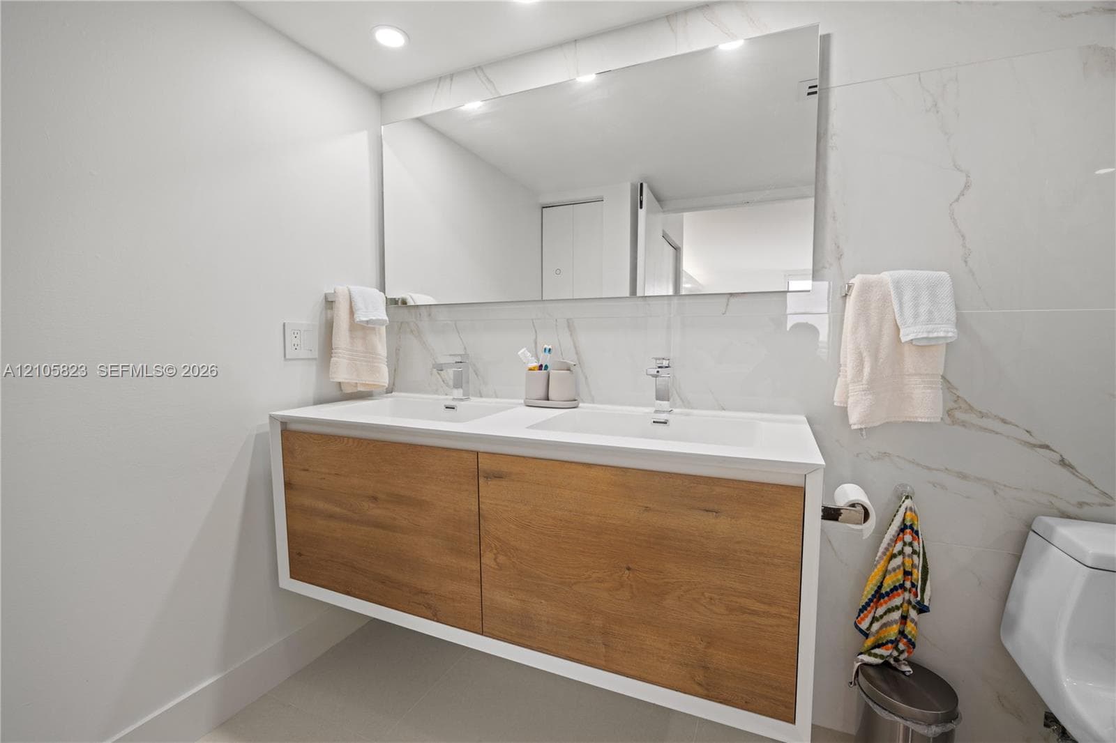 Floating wood-finish double vanity with rectangular integrated sinks, white countertop, polished chrome faucets, full-width wall mirror, and floor-to-ceiling stone-style wall tile