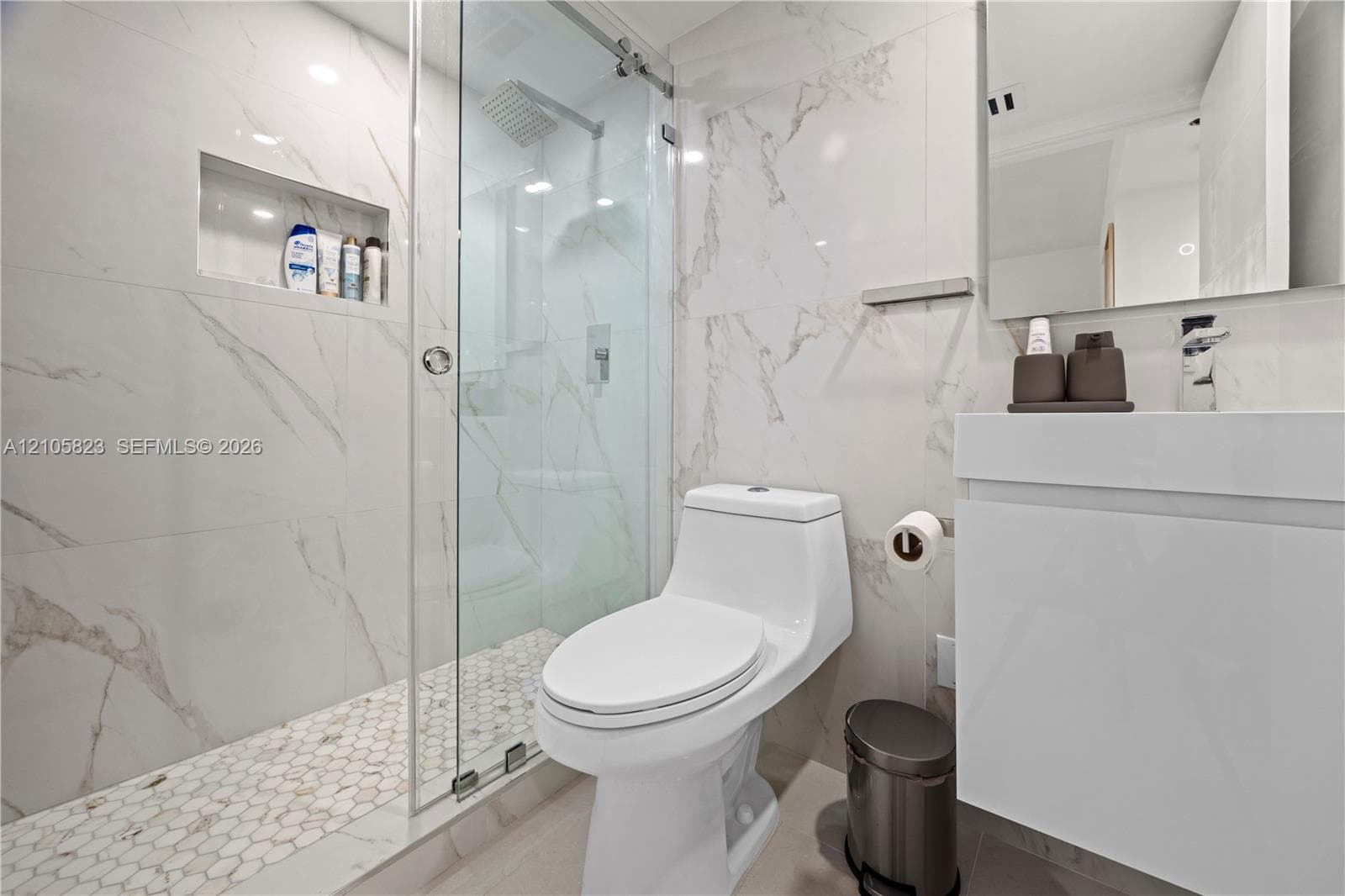 Bathroom featuring wall-to-wall large-format porcelain tile, glass-enclosed shower with integrated shelving, hexagonal floor tiling, and wall-mounted vanity with a minimalist faucet