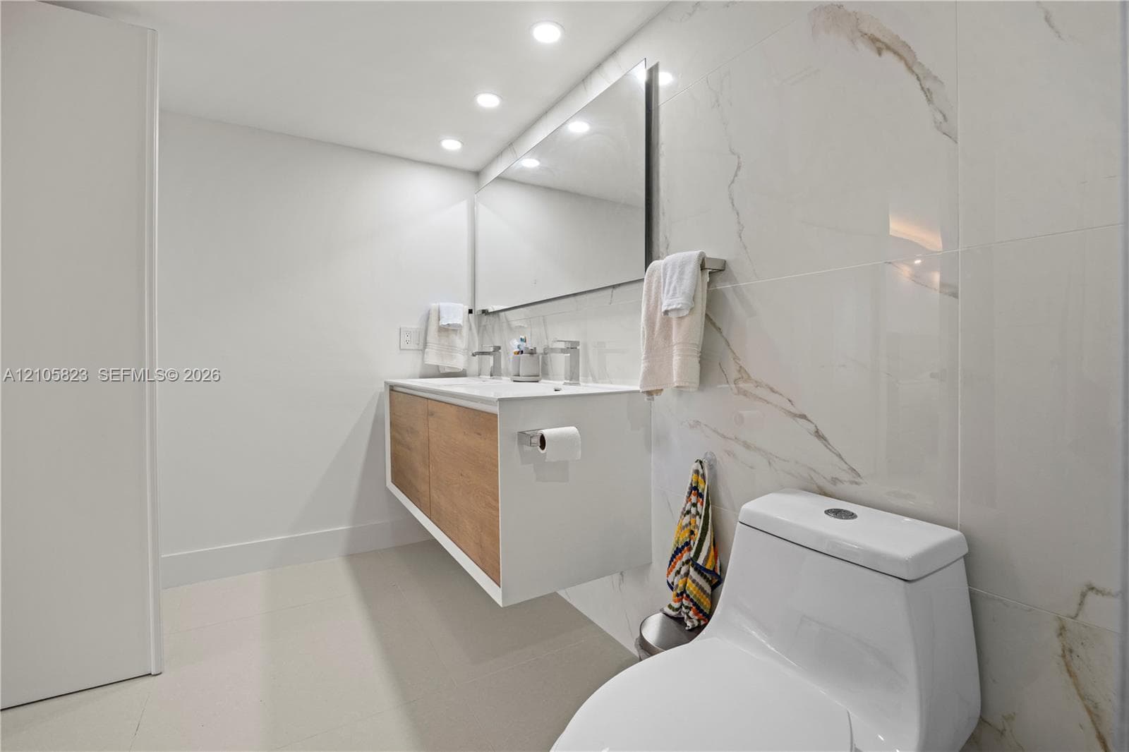 Floating vanity with wood-finish cabinetry, rectangular vessel sink, large wall-mounted mirror, recessed ceiling lighting, and floor-to-ceiling porcelain wall tile