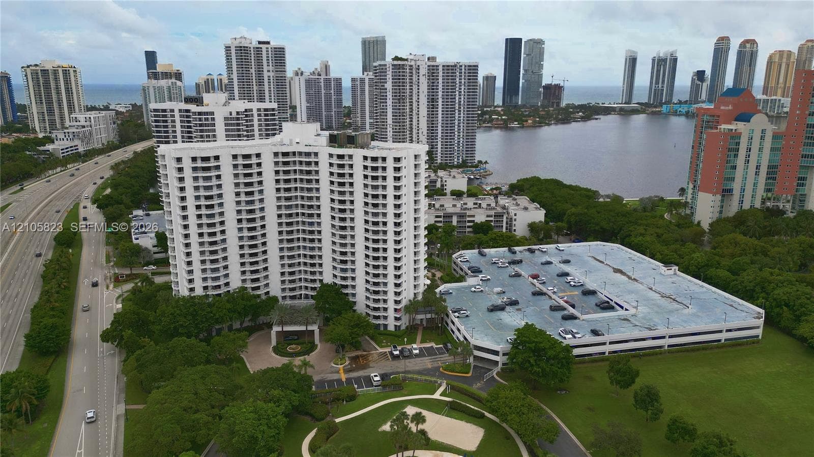 Aerial perspective of a high-rise residential complex featuring a multi-level parking structure, landscaped grounds, and proximity to the coastline