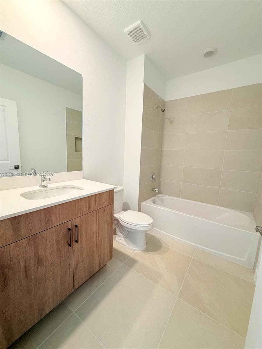 Bathroom featuring a wood-finish vanity with a light-toned countertop, large-format neutral floor tiling, a porcelain-enamel bathtub, and full-height tile shower surround