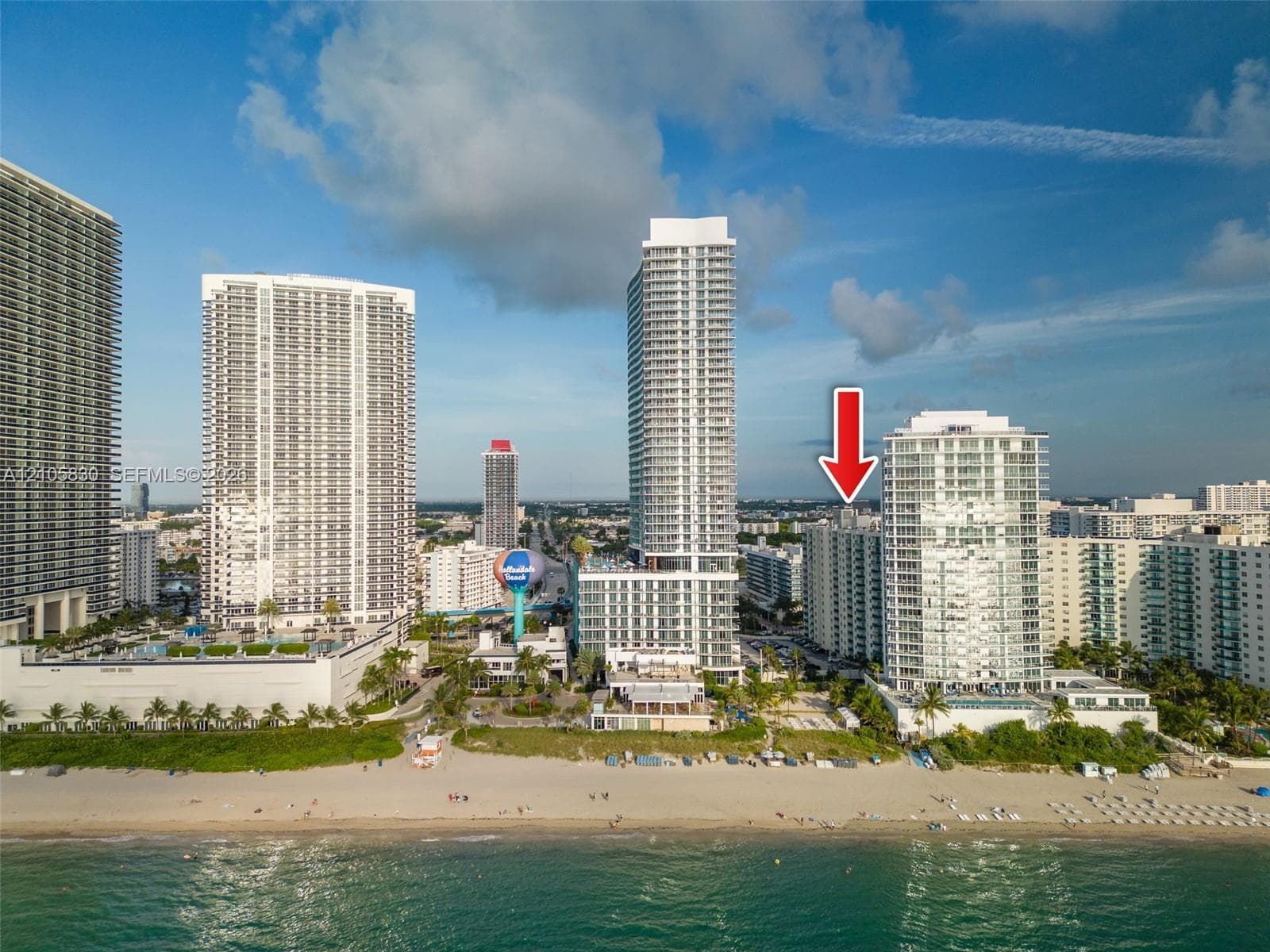 Aerial perspective of a high-rise residential building situated along a coastal shoreline, featuring direct beach access and proximity to regional urban centers