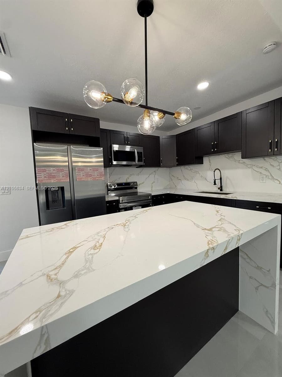 Kitchen featuring an expansive waterfall island with a veined stone-finish countertop, dark cabinetry, stainless steel appliances, and a multi-bulb statement chandelier