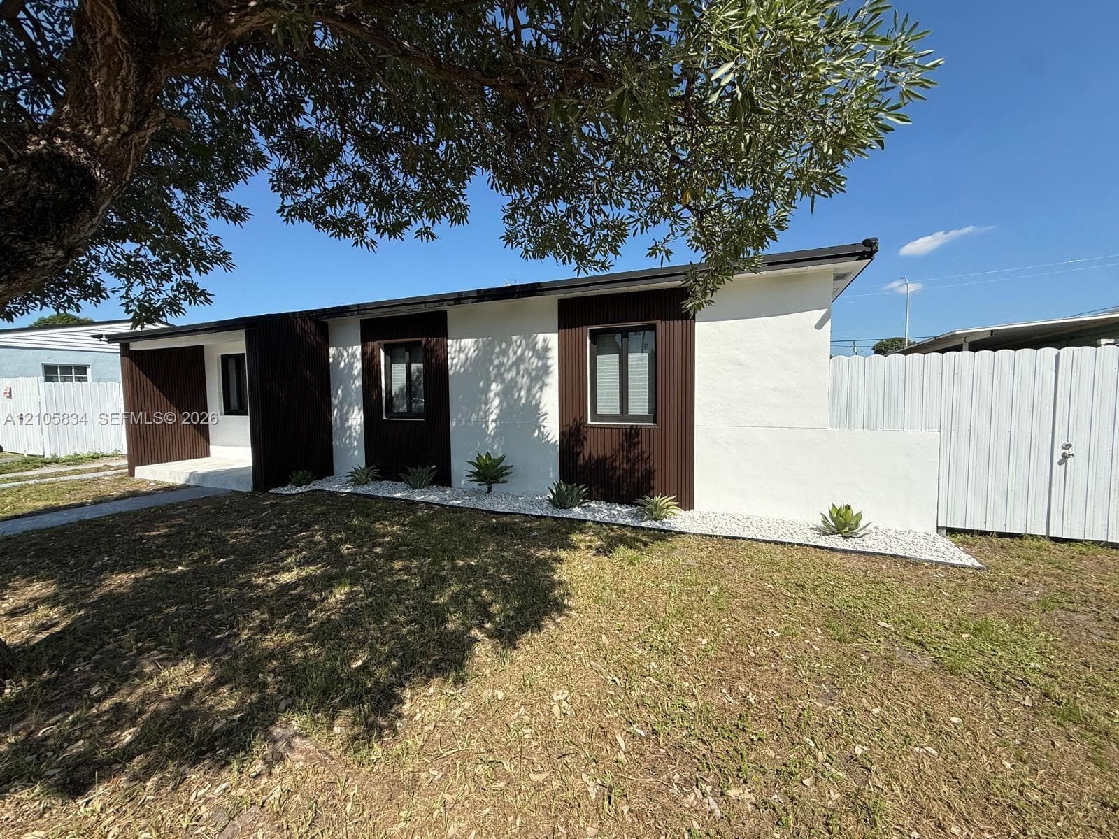 Exterior facade featuring smooth-finish stucco, dark vertical wood-texture siding, low-profile landscaping with white stone ground cover, and a private vinyl fence