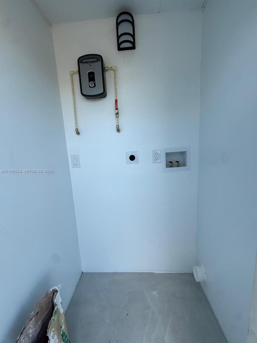 Utility alcove featuring a wall-mounted electric tankless water heater, recessed plumbing connections, an integrated electrical outlet, and a dedicated wall exhaust vent