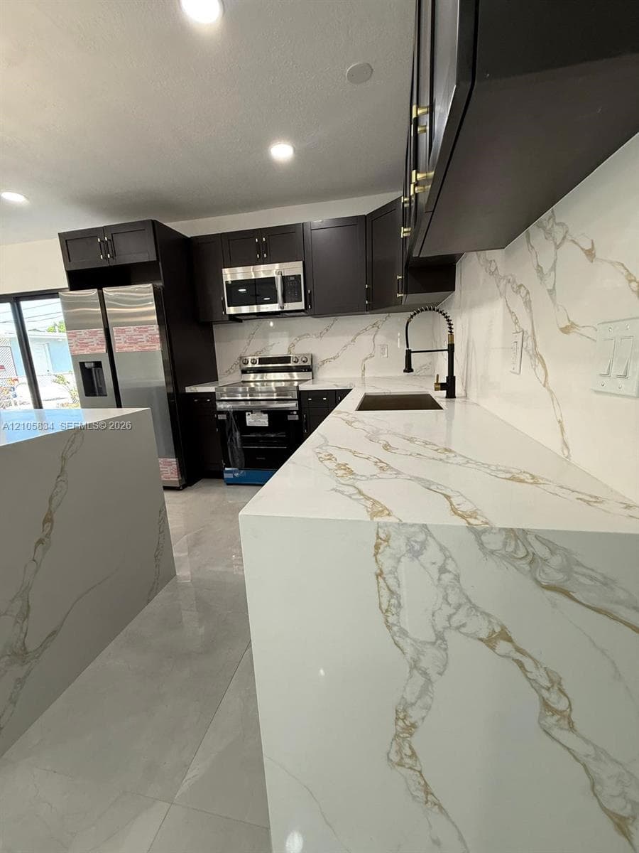 Kitchen featuring dark shaker-style cabinetry, white countertops with gold veining, stainless steel appliances, a pull-down kitchen faucet, and large-format tile flooring