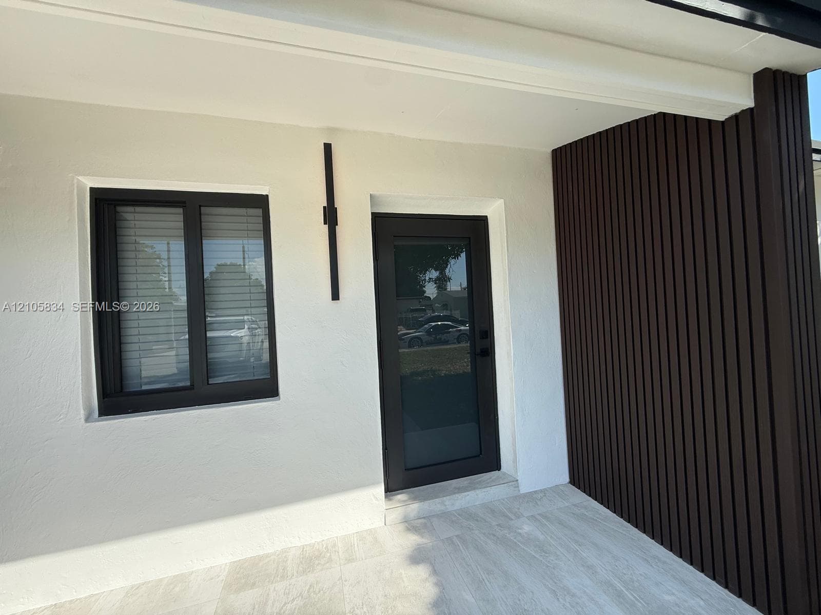 Entryway featuring a smooth white stucco facade, dark vertical wood-slat accent wall, tile flooring, glass-panel exterior door, and a slim black vertical wall-mounted light fixture