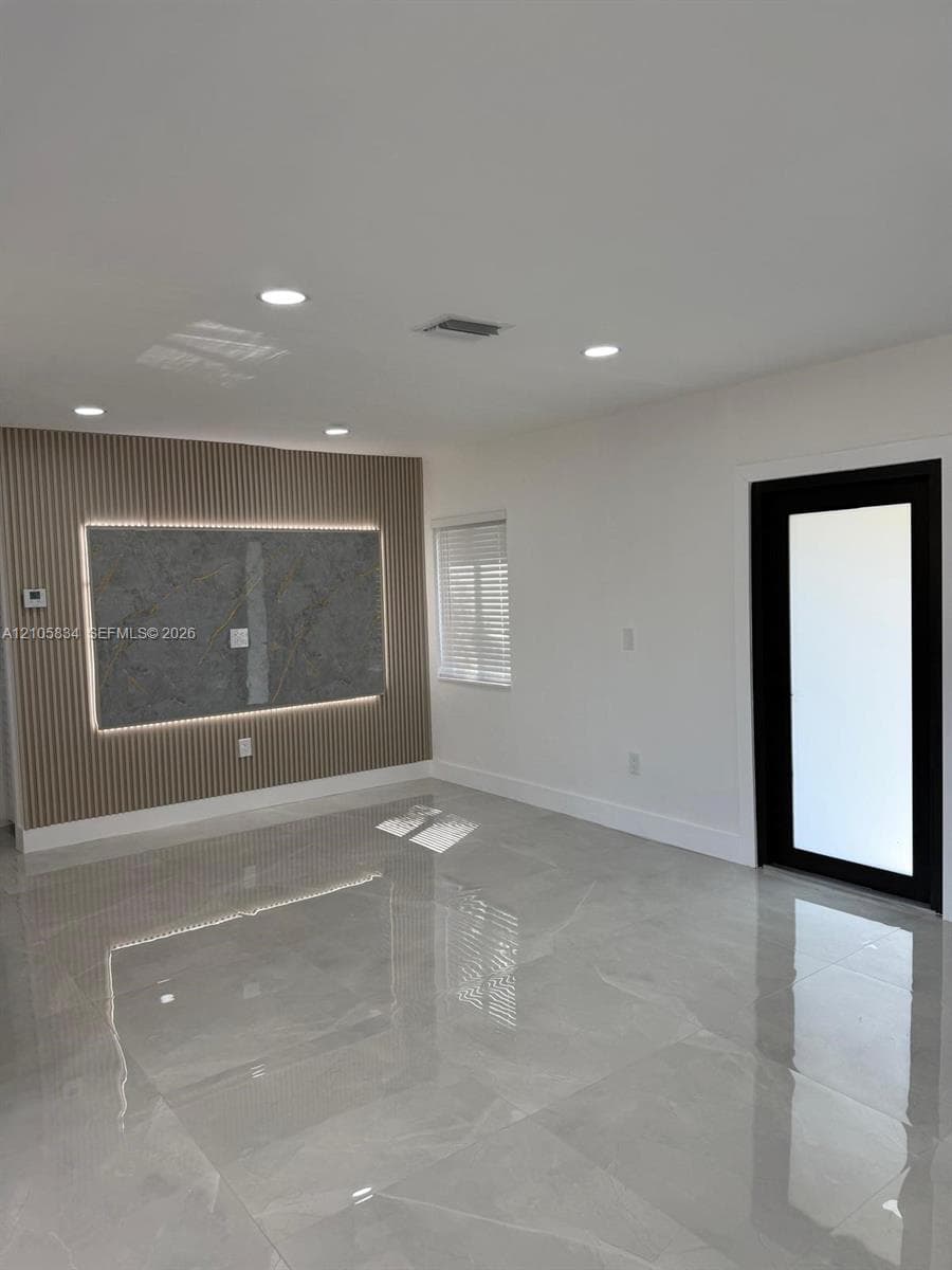 Polished large-format tile flooring, accent wall featuring vertical slat paneling with integrated lighting, centered stone-finish panel, and a black-framed glass door