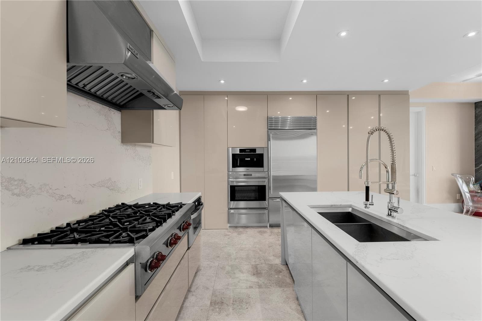 Kitchen featuring flat-panel cabinetry, a stainless steel gas range with a professional-grade hood, stone countertops with matching backsplash, built-in stainless steel appliances, and a central island with a double-basin undermount sink