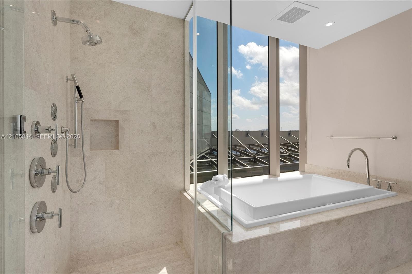 Walk-in shower featuring natural stone tiling, recessed wall niche, and multi-head chrome fixtures, accompanied by a drop-in soaking tub with floor-to-ceiling windows