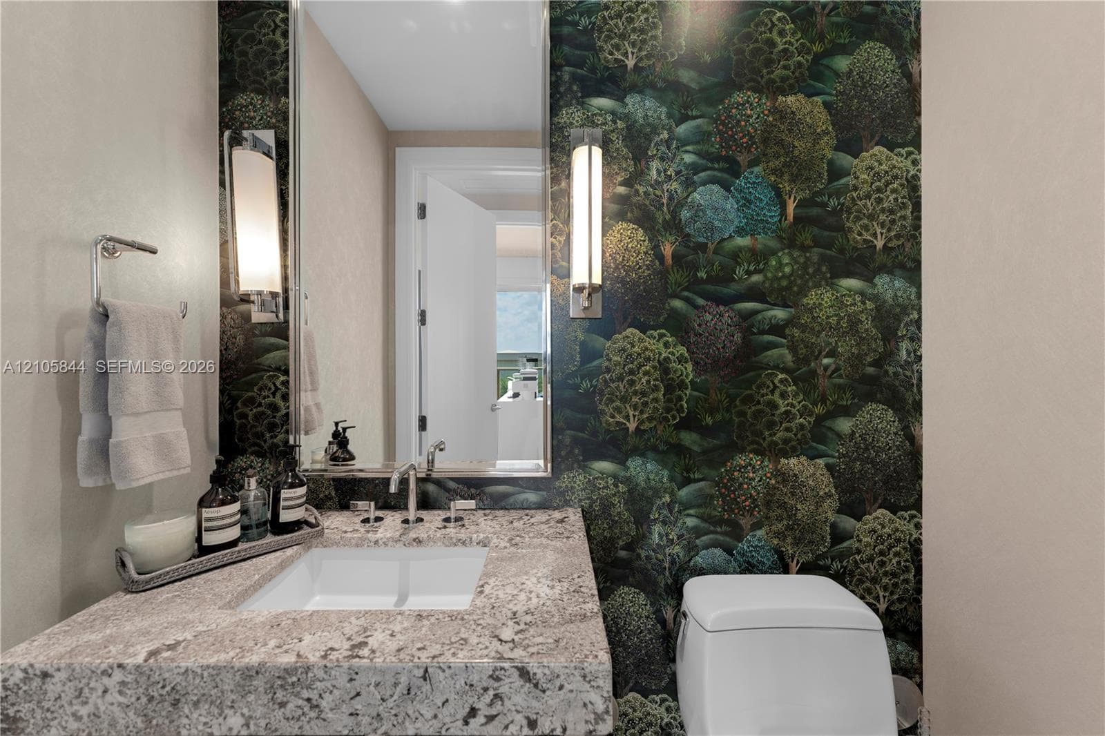 Stone-surfaced vanity with an integrated rectangular sink, wall-mounted metal faucet, floor-to-ceiling forest-motif wallpaper, and dual vertical wall sconces
