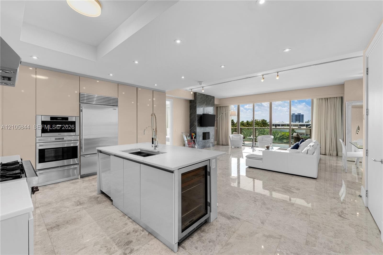 Kitchen featuring high-gloss cabinetry, stone-finish flooring, a central island with an integrated beverage cooler, stainless steel wall-mounted oven and microwave, and floor-to-ceiling windows