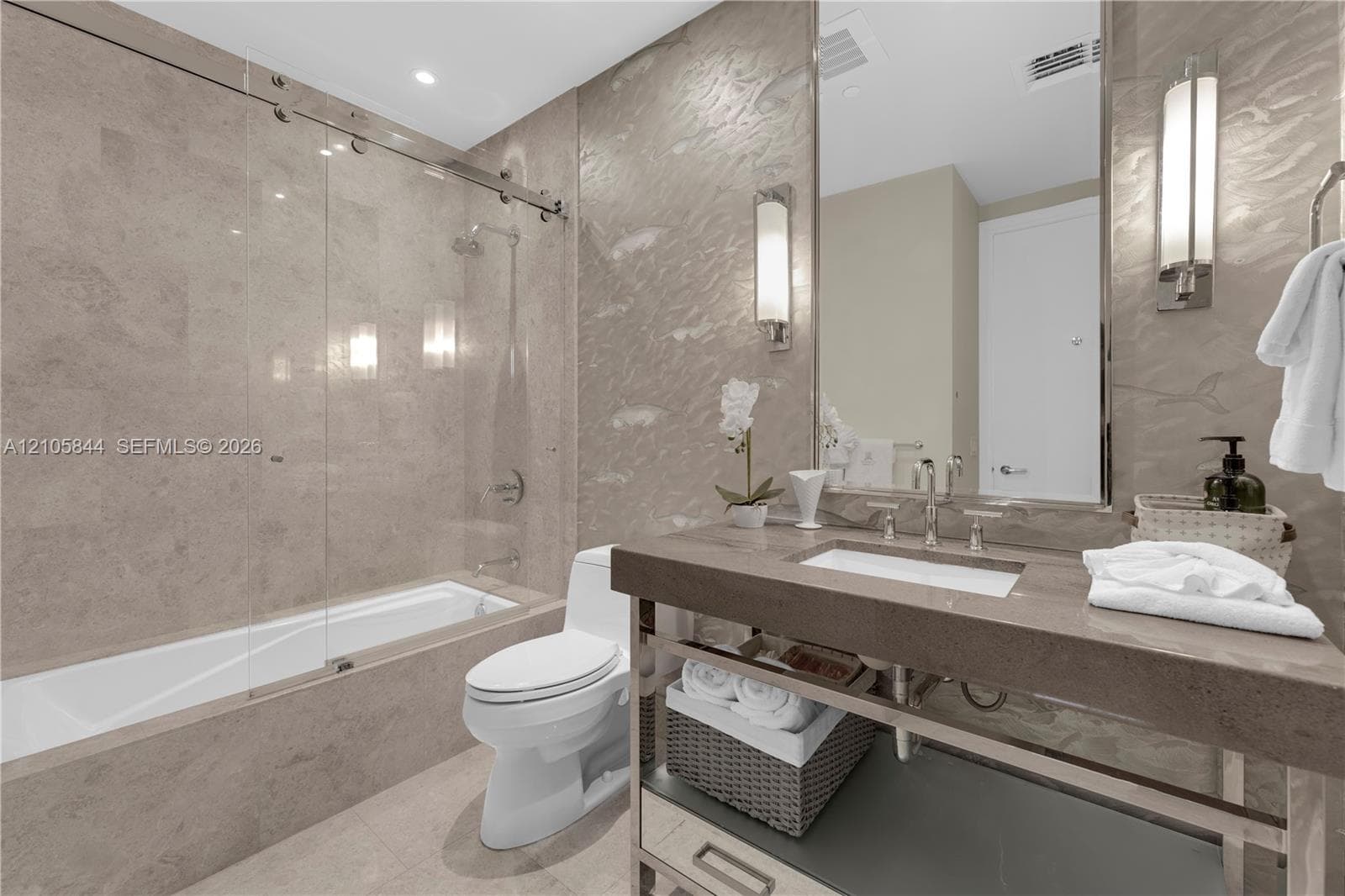 Stone-tiled bathtub enclosure with a frameless glass sliding door, stone-topped vanity with an integrated basin, metal-framed open base, and vertically mounted wall sconces