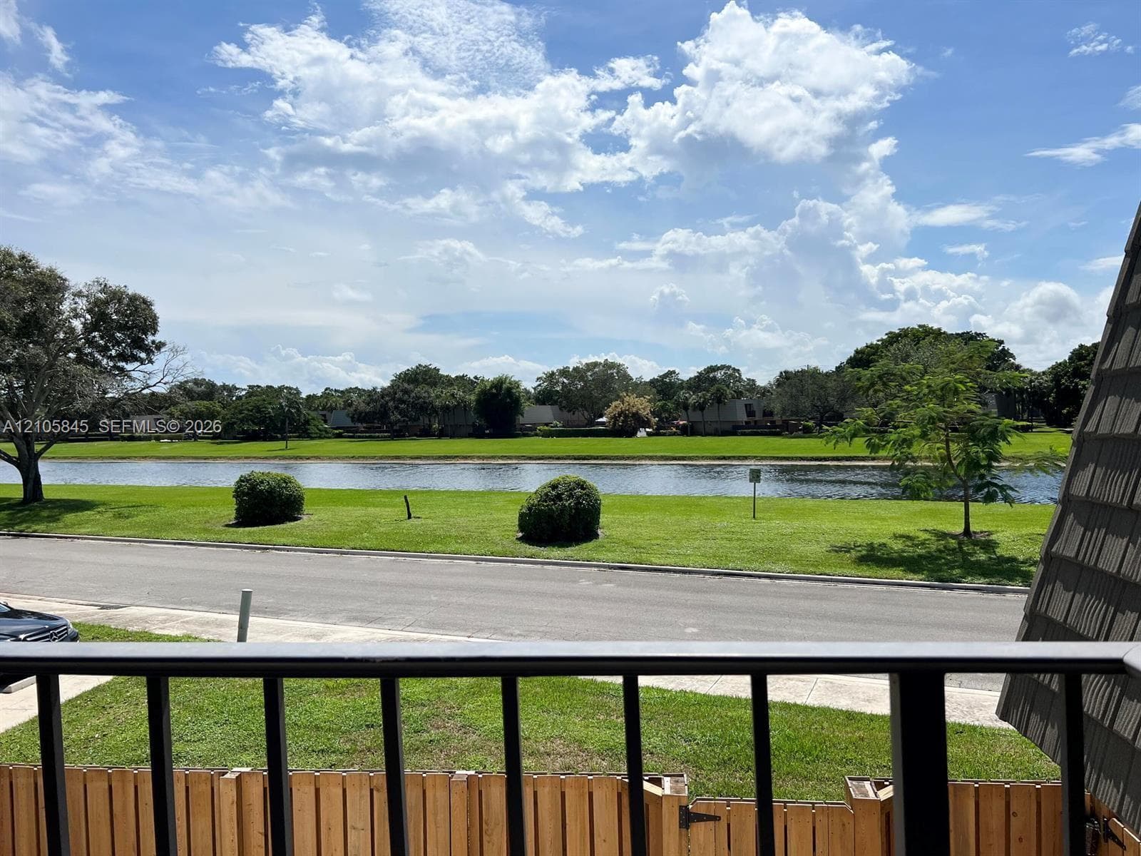 Outdoor balcony featuring a metal railing, overlooking a water feature, manicured landscaping, and asphalt paving