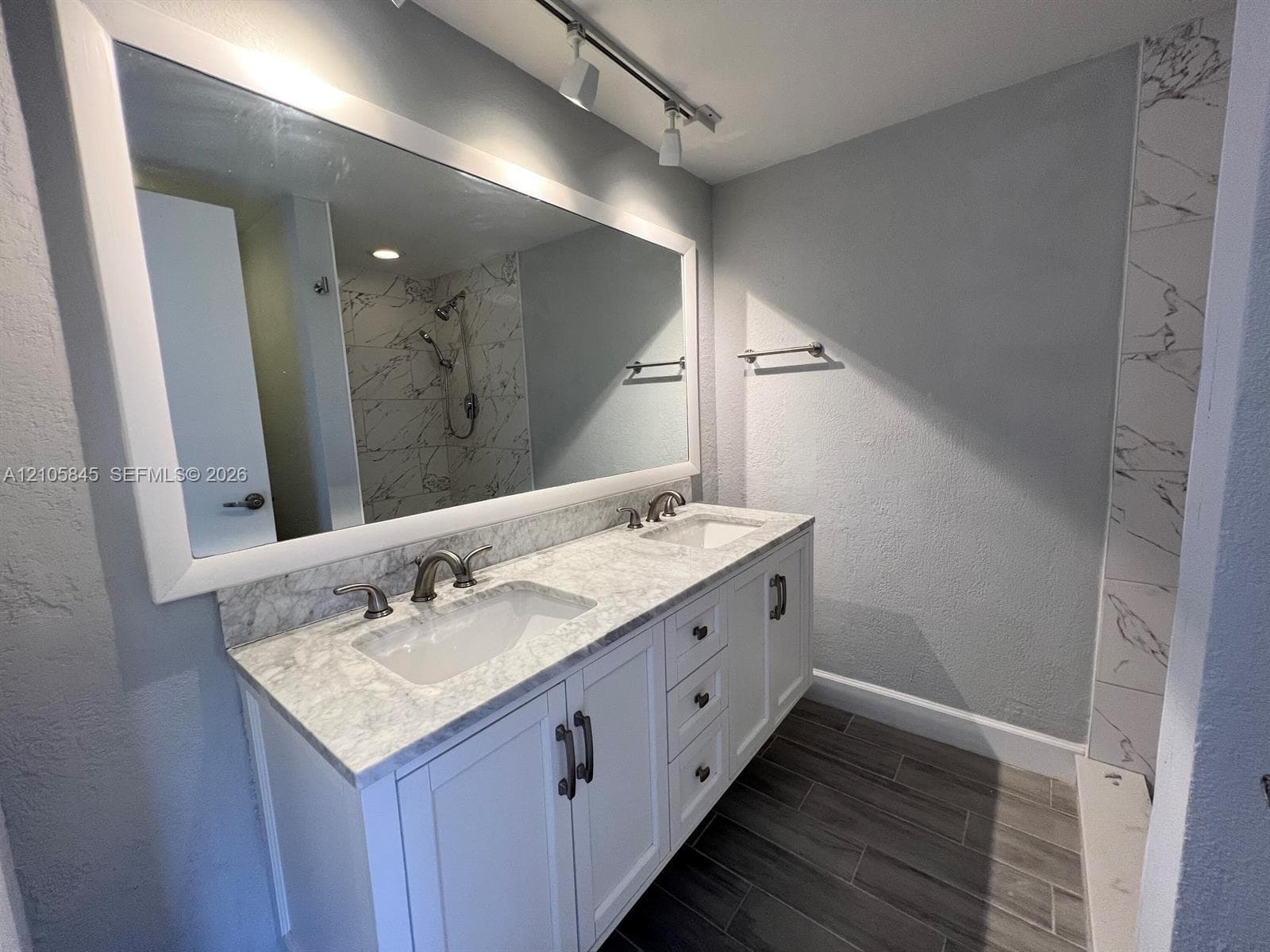 Double vanity with white cabinetry, stone-surface countertop, dual undermount sinks, wide-frame mirror, and wood-finish flooring