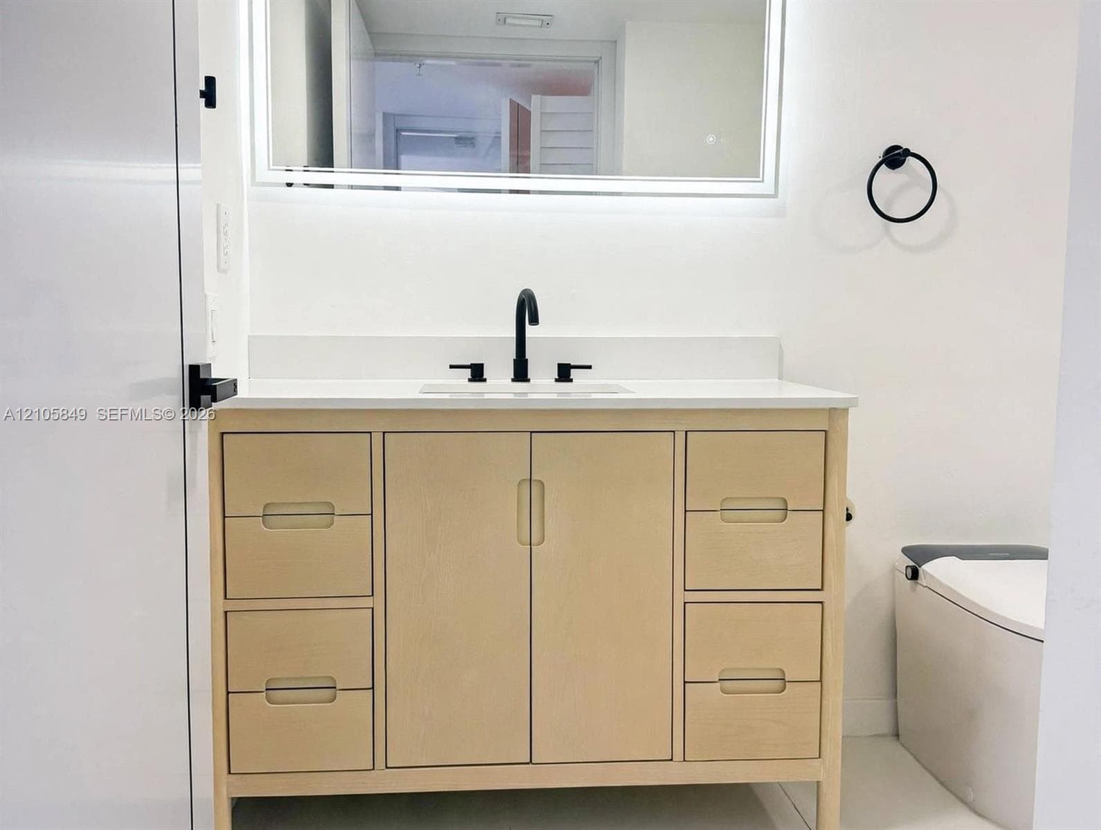 Bathroom featuring a light wood vanity with a white countertop, wall-mounted LED-backlit mirror, matte black fixtures, and a glass-enclosed tub with patterned tile detailing