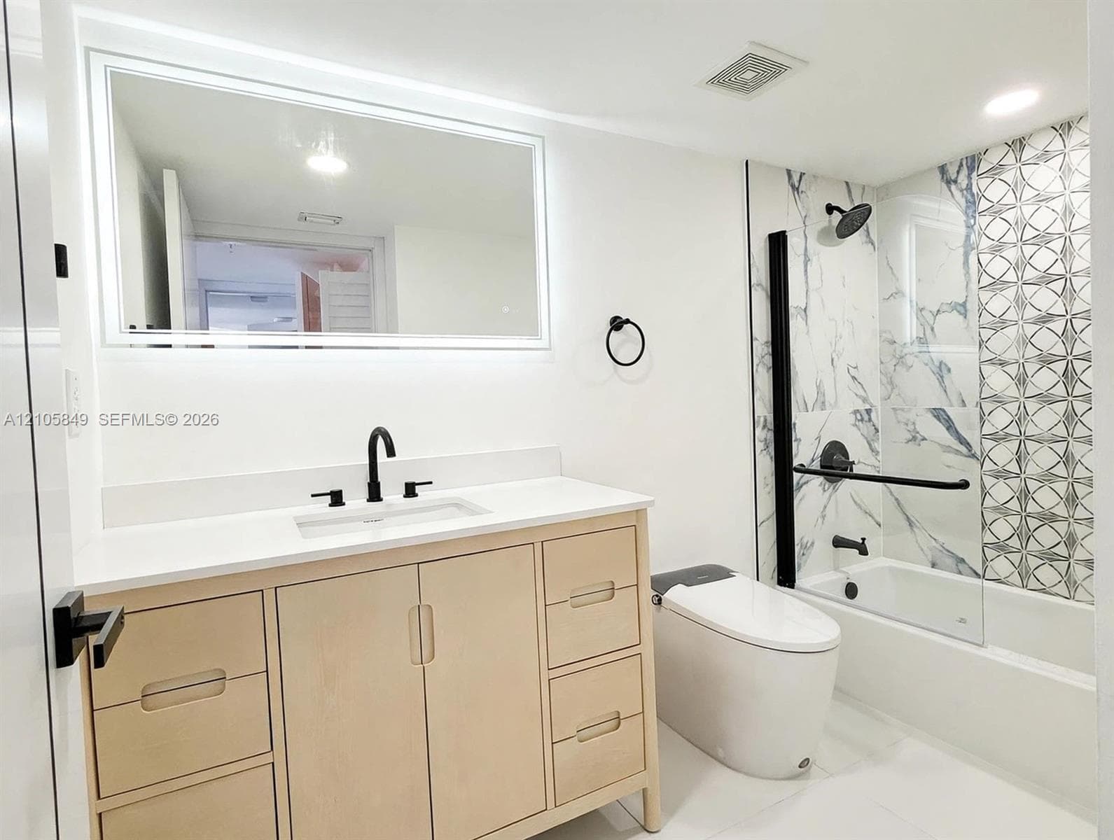 Bathroom featuring a light wood-finish vanity, large-format tile flooring, and a glass-enclosed shower with decorative accent tile, stone-patterned wall panels, and matte black fixtures