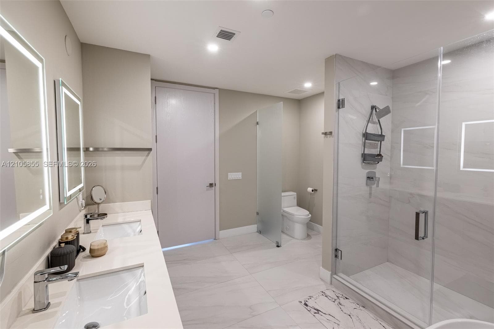 Dual-sink vanity with a solid-surface countertop and integrated lighting, walk-in shower with stone-style wall tile and recessed shelving, and a glass-enclosed water closet