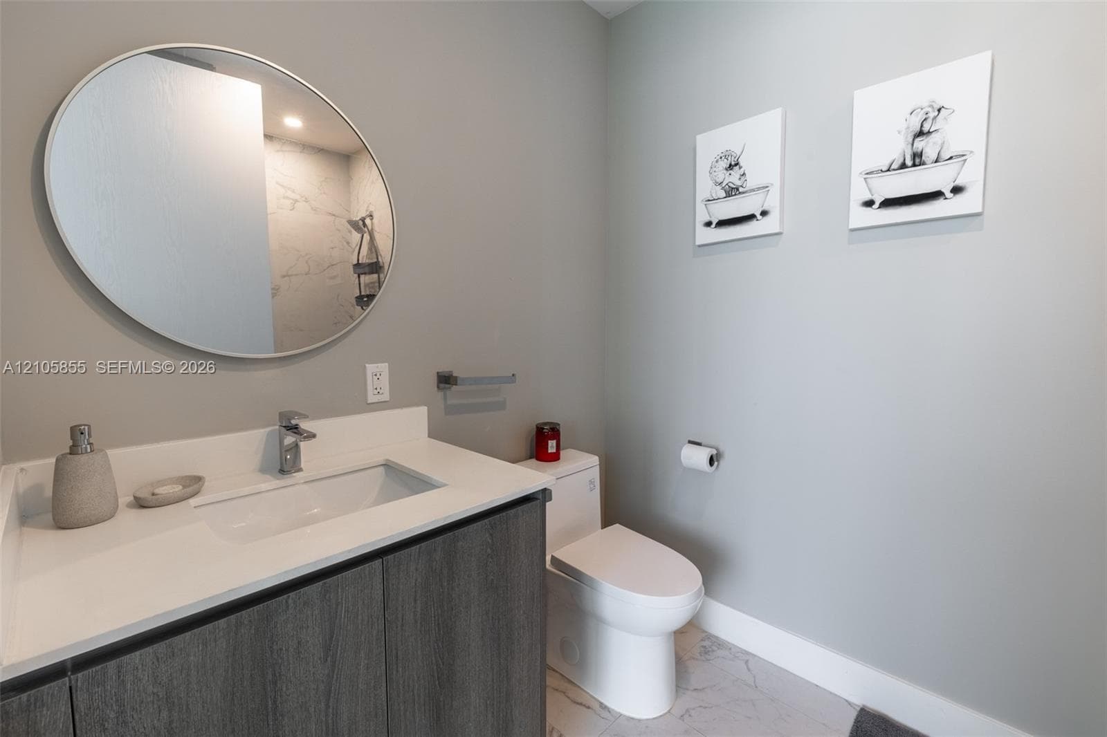 Bathroom featuring a dark wood-finish vanity with a white countertop and rectangular integrated sink, circular wall-mounted mirror, polished chrome faucet, and stone-finish flooring