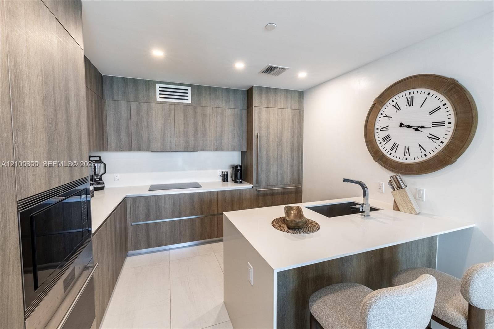 Kitchen featuring flat-panel wood-finish cabinetry, white countertops, a central island with a built-in sink, recessed lighting, and integrated stainless steel appliances