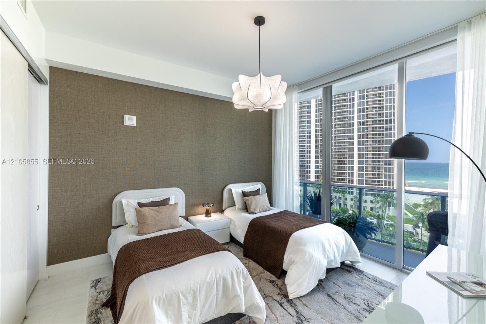 Textured accent wall, floor-to-ceiling windows, sculptural pendant lighting, and a glass-railed balcony with ocean and city scenery