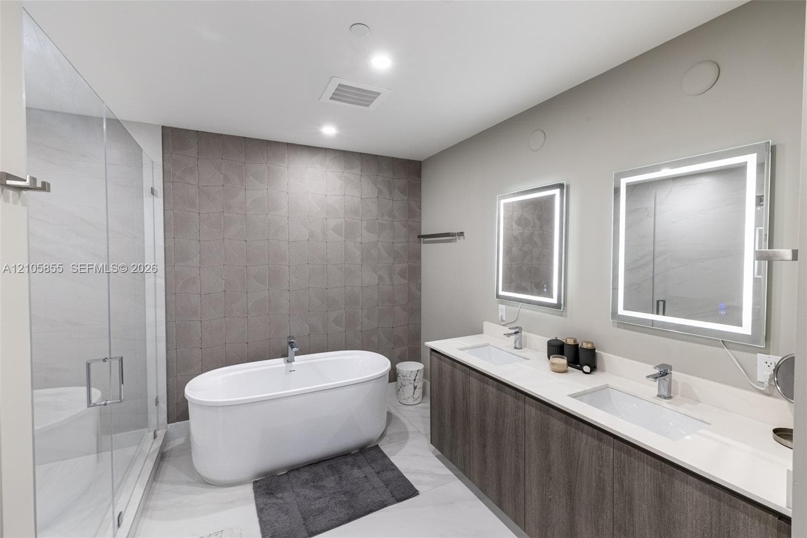 Freestanding soaking tub with a floor-mounted faucet, walk-in glass shower, dual-sink vanity with light-toned stone countertop, wood-finish cabinetry, and LED-backlit mirrors