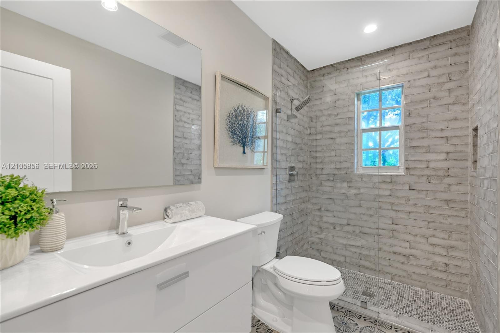 Bathroom featuring a white vanity with integrated basin, large frameless mirror, walk-in shower with textured tile surround, pebble-style floor tile, and chrome fixtures