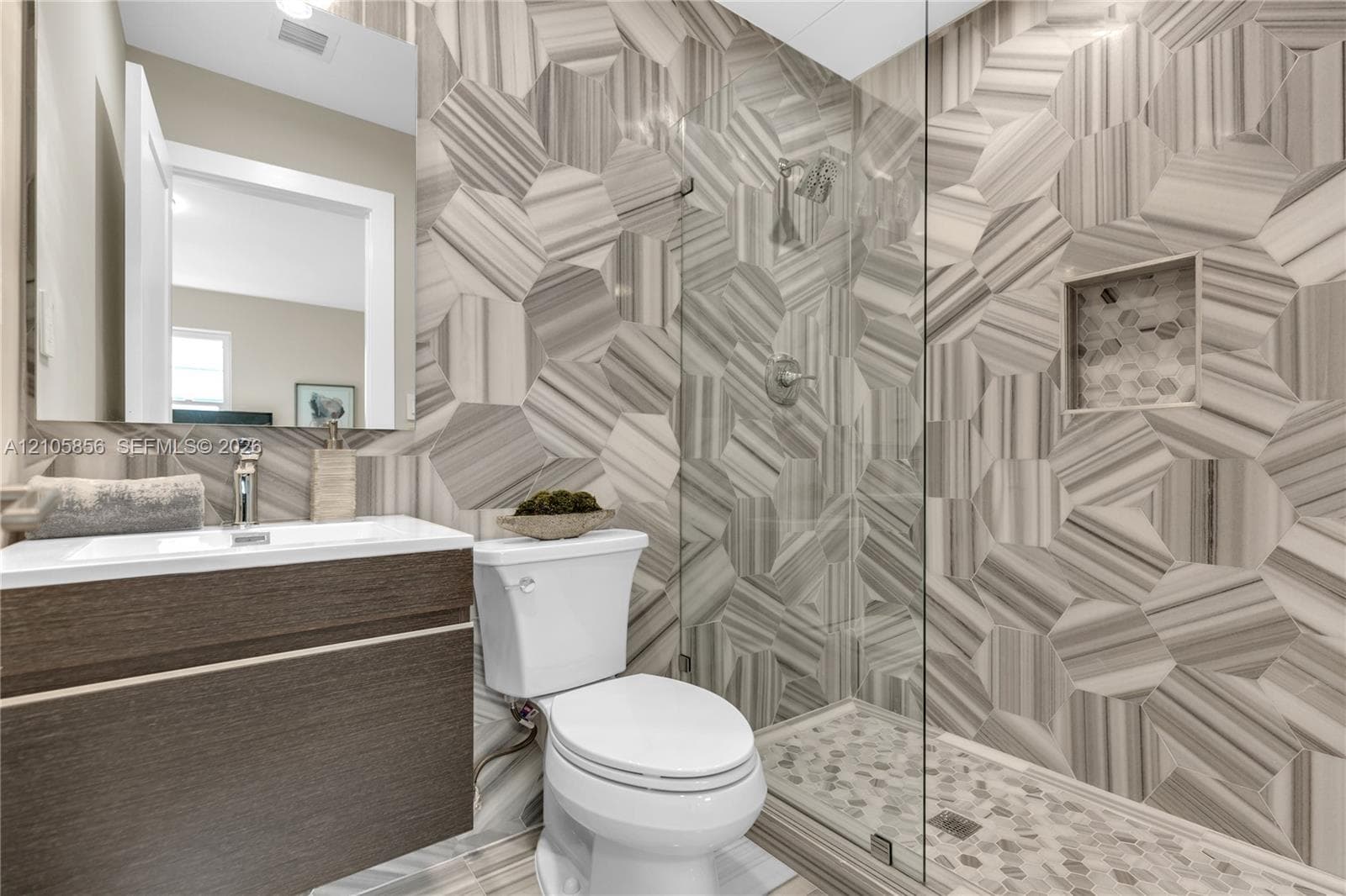 Floating wood-finish vanity with an integrated sink, floor-to-ceiling hexagonal tile shower surround, glass-enclosed shower stall, built-in shower niche, and pebble-style stone floor tiling