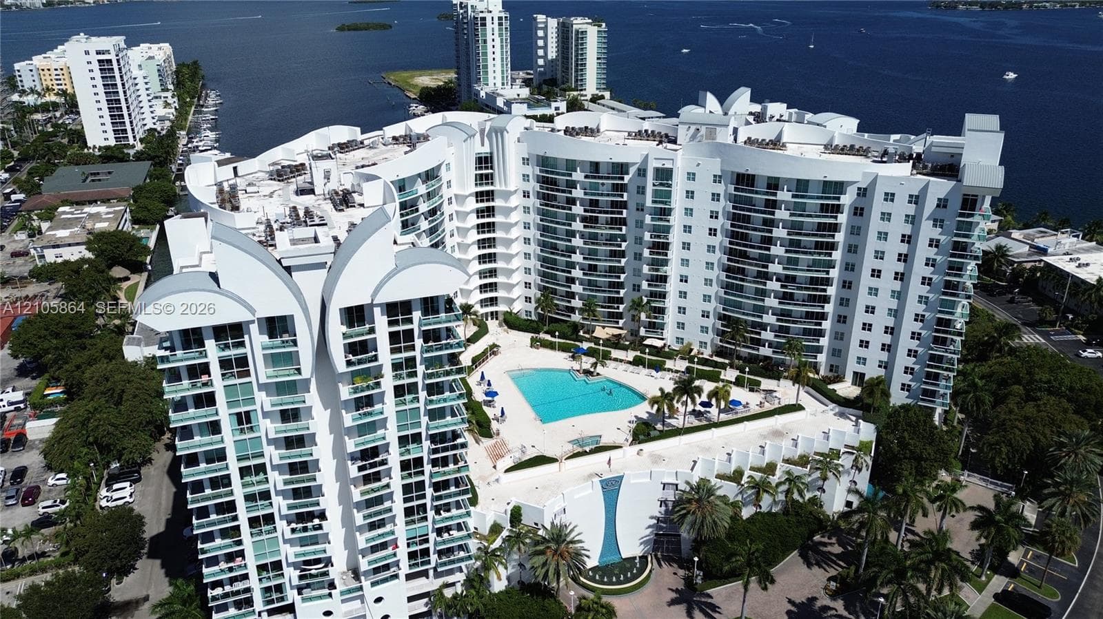 High-rise waterfront property featuring a large swimming pool, expansive paved deck, glass-railed private balconies, and mature palm landscaping