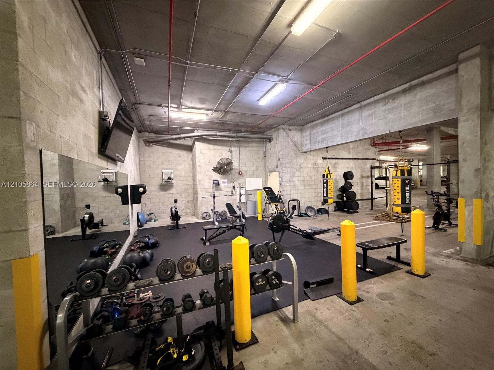 Fitness area featuring exposed concrete ceiling, industrial-style lighting, large wall-mounted mirror, and impact-resistant rubber flooring