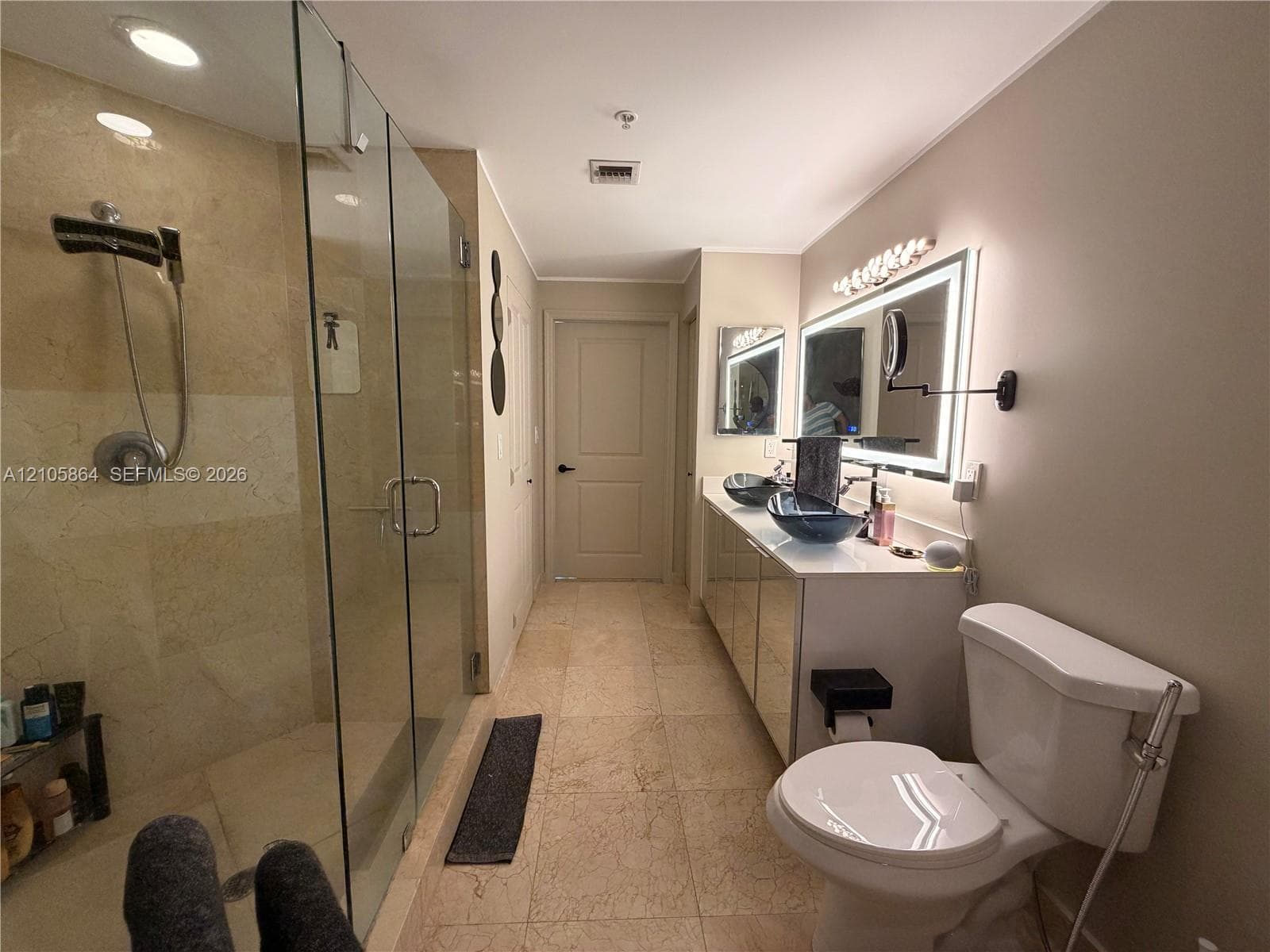 Bathroom featuring stone-tile flooring, glass-enclosed shower with stone wall surfacing, dual vanity with vessel sinks, backlit wall-mounted mirror, and wall-mounted articulating vanity mirror