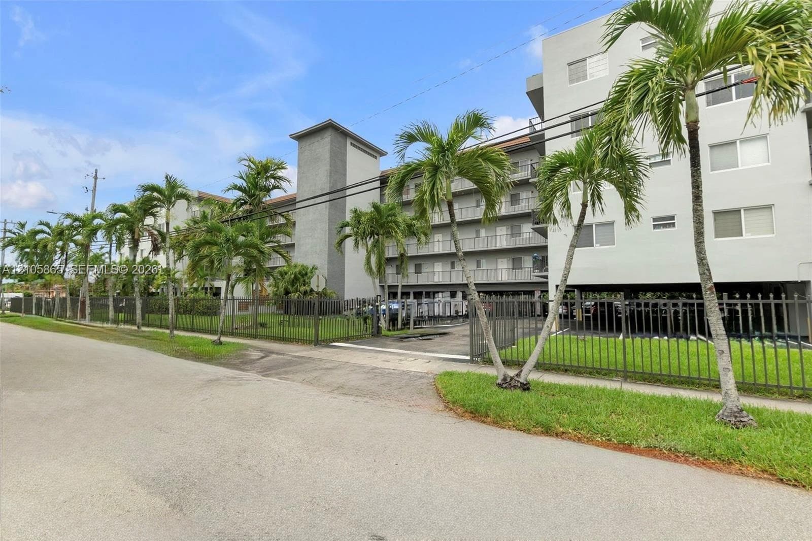 Multi-story building exterior featuring a neutral stucco facade, grey vertical accent detailing, metal balcony railings, and a perimeter metal fence