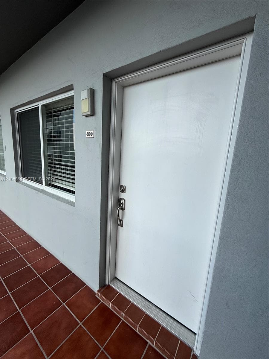 Entryway featuring a solid white door with a silver-finish handle set, terracotta-toned tile flooring, and exterior wall-mounted lighting