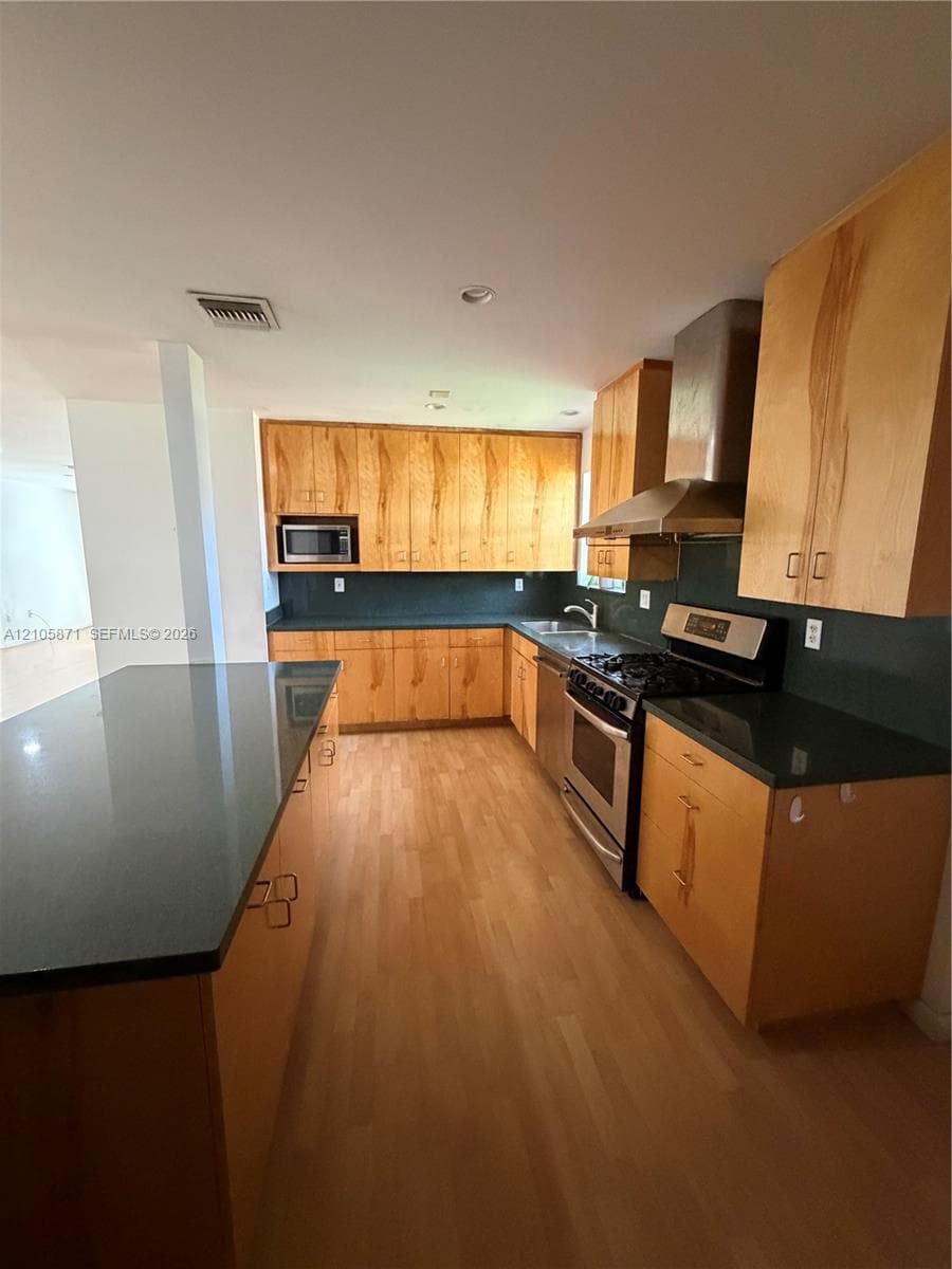 Kitchen featuring light wood-finish cabinetry, dark solid-surface countertops, wood-finish flooring, stainless steel range with matching vent hood, and a built-in microwave