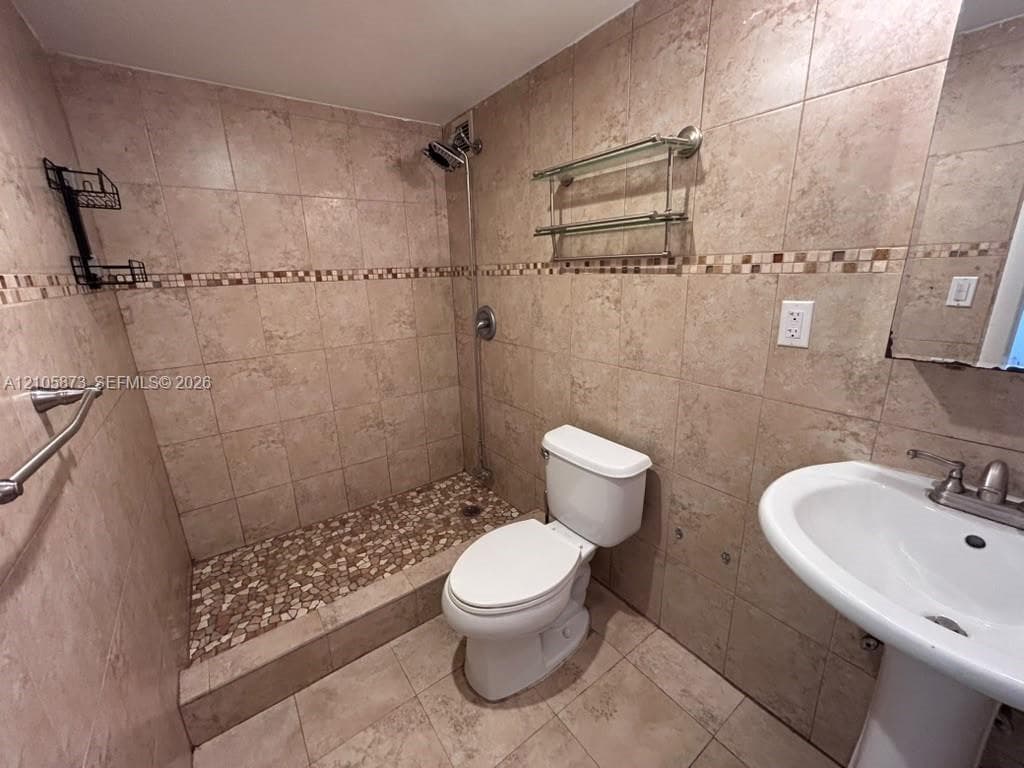 Full-height natural stone tiling, shower stall with pebble-finish flooring, pedestal sink, chrome-finished plumbing fixtures, and integrated glass shelving