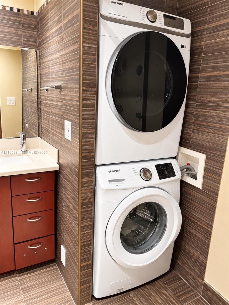 Stacked front-load laundry unit set against floor-to-ceiling textured tile wall, paired with a vanity featuring a solid surface countertop, rectangular undermount sink, and cherry-finish cabinetry