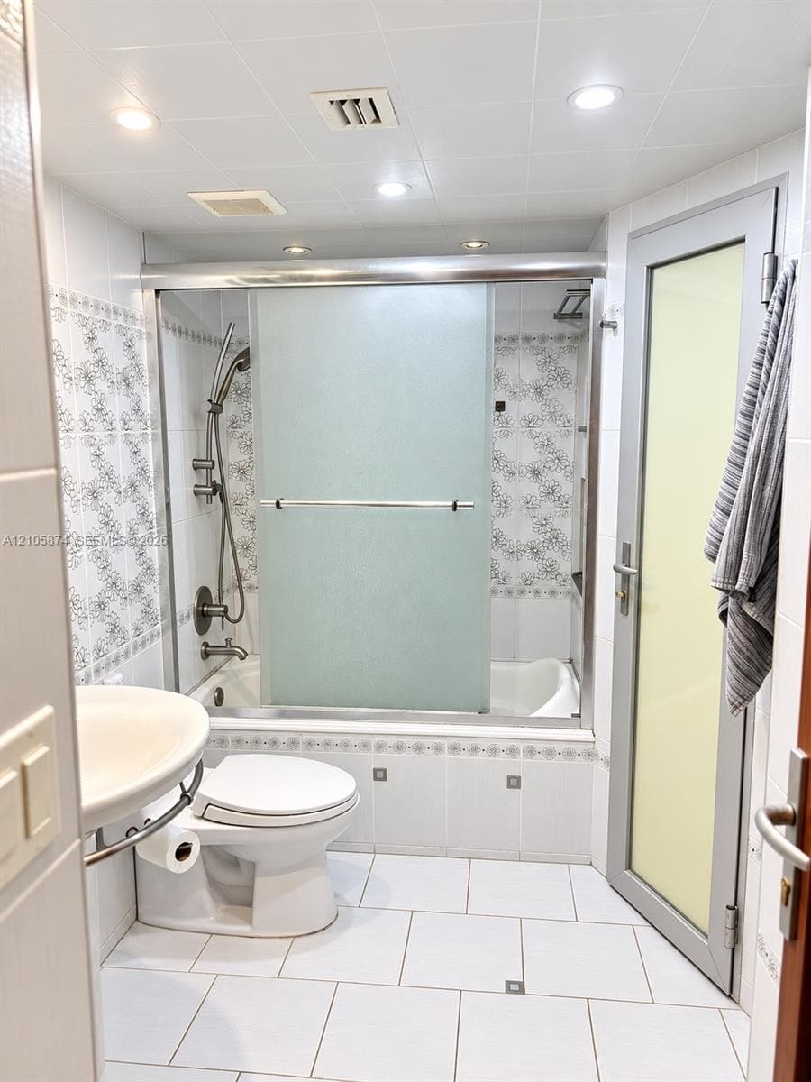 Full-bath featuring a bathtub with a sliding frosted-glass enclosure, wall-mounted shower hardware, decorative floral tile backsplash, ceramic tile flooring, and a pedestal sink