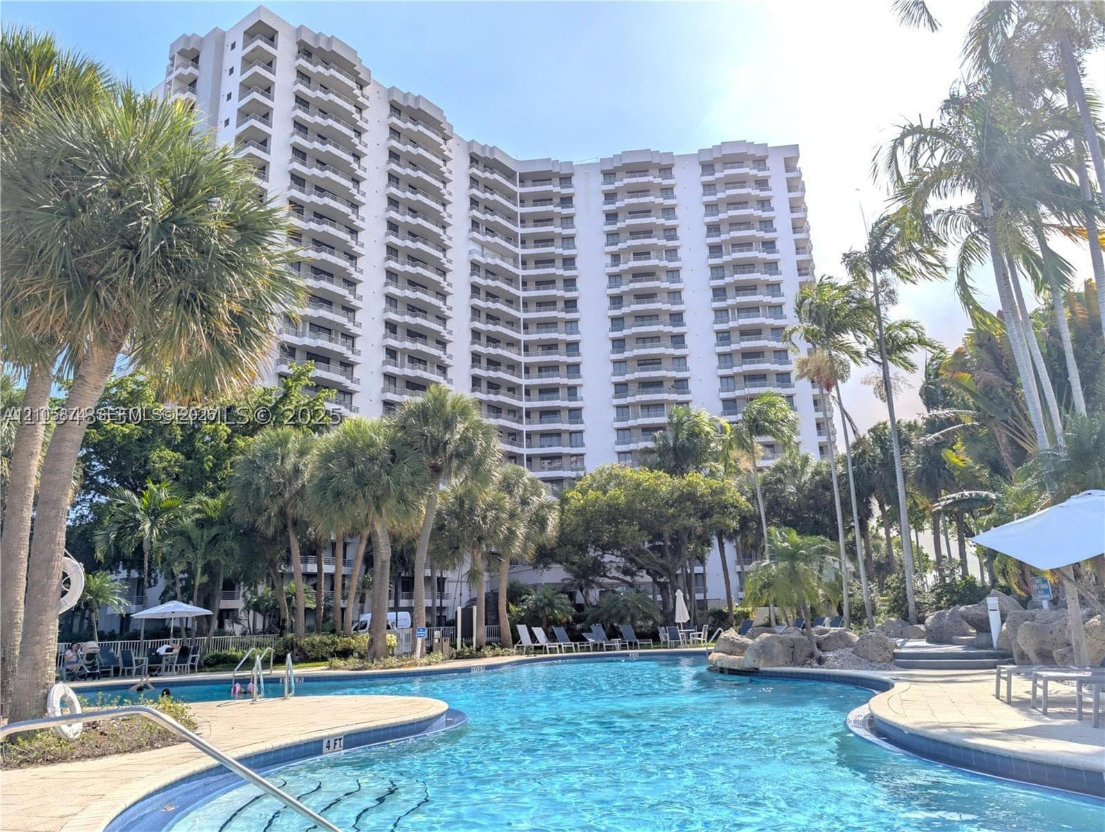 High-rise exterior featuring private balconies and an expansive swimming pool with hardscaped pool deck, rock features, and mature palm trees