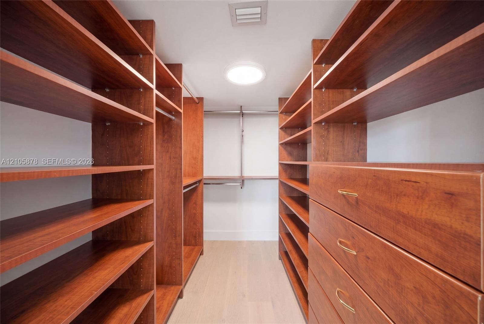 Walk-in storage space featuring extensive custom wood-finish shelving, integrated clothing rods, built-in dresser drawers with metallic pulls, and light-toned flooring