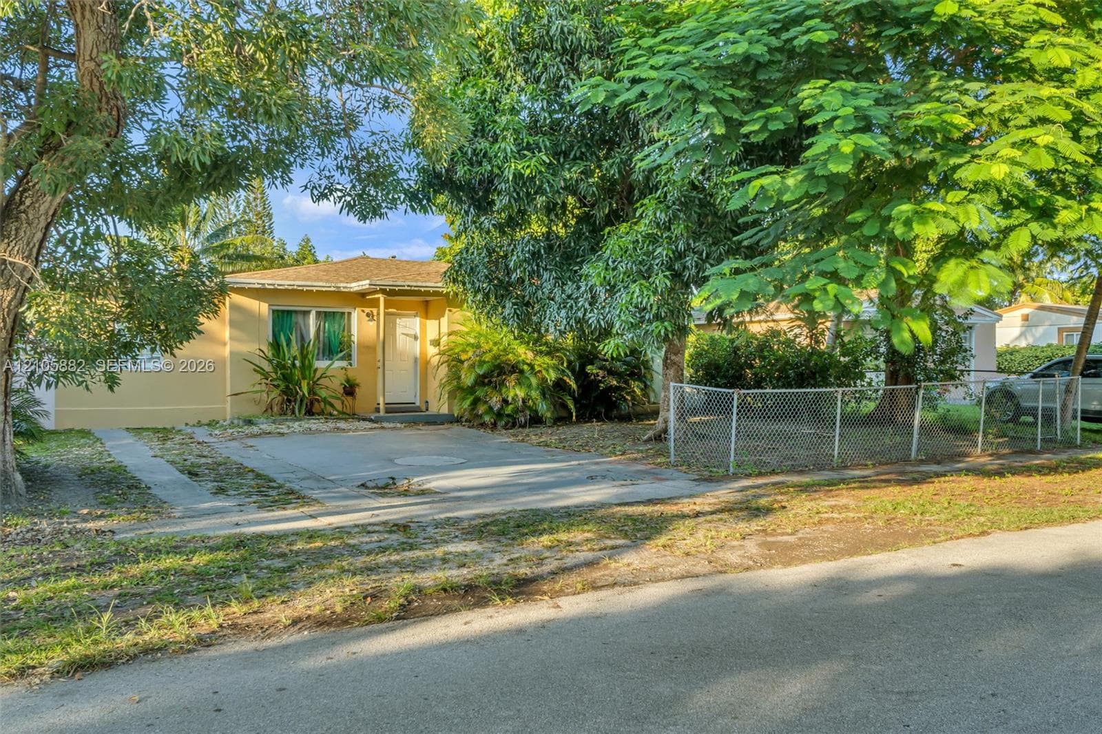 Single-story residence featuring a concrete driveway, mature canopy trees, and perimeter chain-link fencing